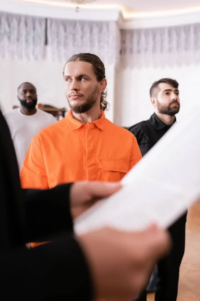 Accused man in orange jail uniform standing near bailiff and prosecutor with paper on blurred foreground — Stock Photo