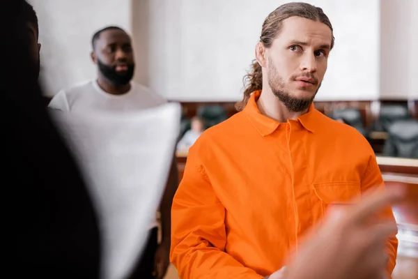 Accused man in jail uniform looking at blurred prosecutor pointing with finger in courtroom — Stock Photo