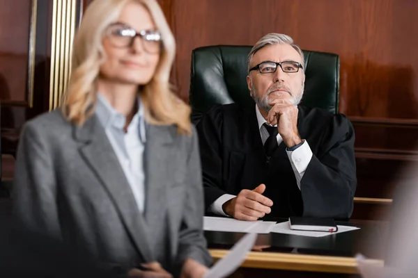 Grey-haired judge in robe and eyeglasses near blurred advocate in courtroom — Stock Photo