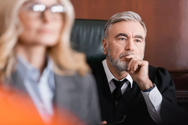 Juge principal tenant la main près du visage tout en pensant avocat proche sur le premier plan flou — Photo de stock