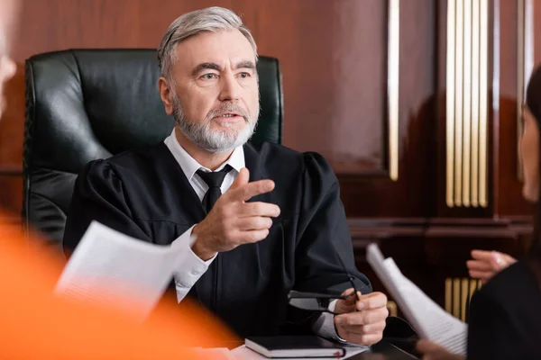 Oberster Richter zeigt mit erhobenem Zeigefinger nahe verschwommenem Staatsanwalt mit Papieren vor Gericht — Stockfoto