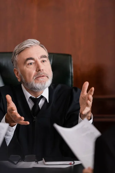 Juiz sênior apontando com as mãos enquanto conversa com promotor desfocado com processo — Fotografia de Stock