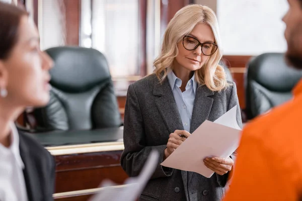 Middle aged advocate looking at documents near prosecutor and accused man on blurred foreground — Stock Photo