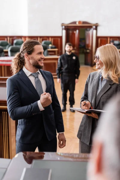 Оправданный бизнесмен, показывающий успешный жест рядом с улыбающимся адвокатом и размытым судьей в суде — стоковое фото