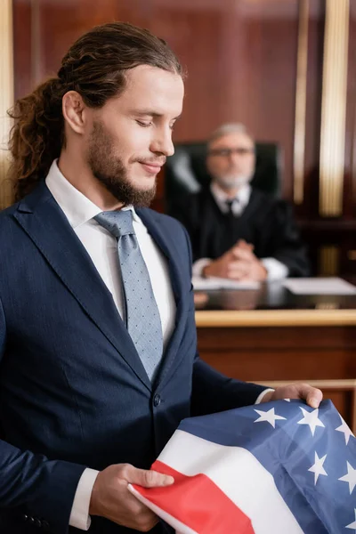 Позитивный адвокат держит флаг США в зале суда рядом со старшим судьей на размытом фоне — стоковое фото