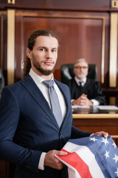 Advogado olhando para a câmera enquanto segurando bandeira dos EUA perto de juiz desfocado no tribunal — Fotografia de Stock
