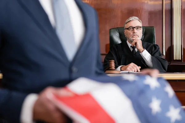 Grey-haired judge looking at camera in court while attorney holding usa flag on blurred foreground — Stock Photo