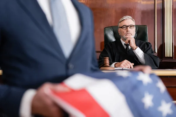 Grauhaarige Richterin blickt vor Gericht in die Kamera, während ihr Anwalt eine US-Flagge im verschwommenen Vordergrund hält — Stockfoto