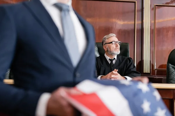 Juez superior mirando hacia otro lado en la sala del tribunal cerca de abogado con bandera de EE.UU. en primer plano borrosa - foto de stock