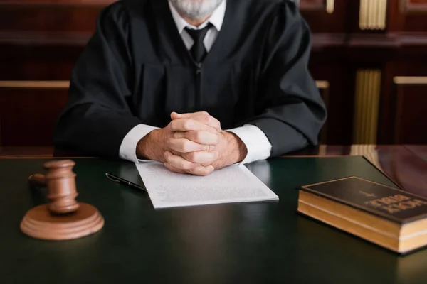 Vue partielle du juge assis les mains serrées près de la bible, du marteau et du procès sur le bureau — Photo de stock