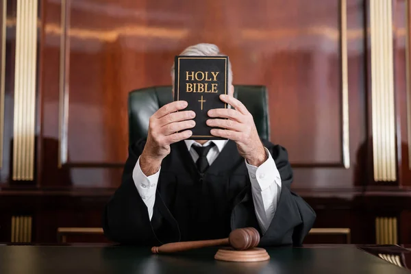 Judge in mantle obscuring face with bible while sitting near gavel in courtroom — Stock Photo