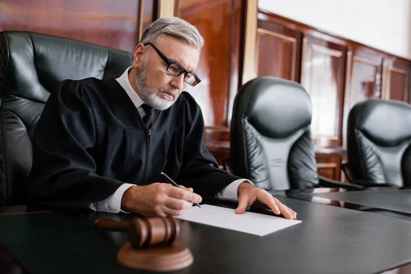 Juiz sênior em roupão e óculos segurando caneta perto de papel e martelo borrado — Fotografia de Stock
