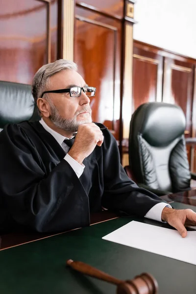 Старший судья в халате и очках сидит с ручкой во время судебного разбирательства — стоковое фото