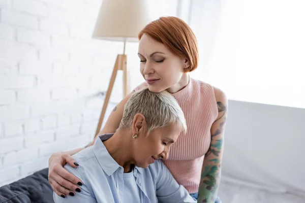 Tattooed lesbian woman with closed eyes hugging smiling girlfriend at home — Stock Photo