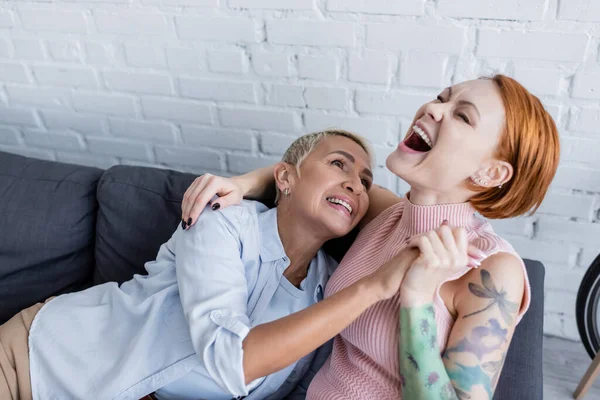 Alegre mujer riendo cerca lesbiana novia en sofá en casa - foto de stock