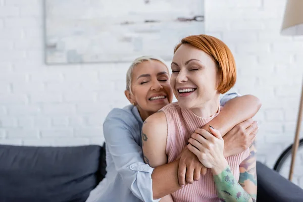 Gioiosa donna lesbica con gli occhi chiusi abbracciando la fidanzata tatuata a casa — Foto stock