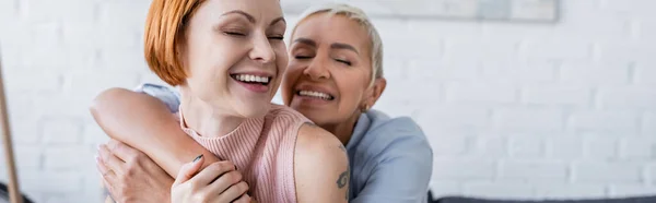 Mujer lesbiana feliz con los ojos cerrados abrazando novia lesbiana en casa, pancarta - foto de stock