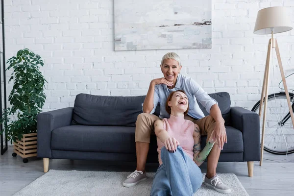 Happy lesbian woman smiling at camera near girlfriend laughing on floor in living room — Stock Photo