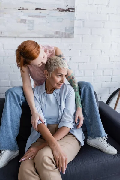 Tattooed woman hugging lesbian girlfriend looking away on couch at home — Stock Photo