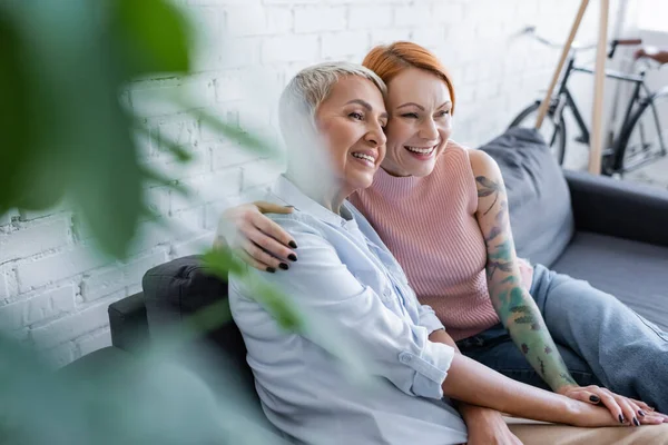 Glückliche lesbische Frauen, die zu Hause auf dem Sofa auf verschwommenem Vordergrund sitzen — Stockfoto