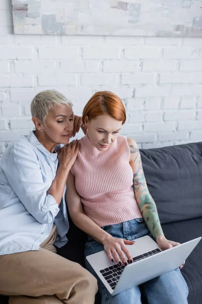 Lesbienne femme assise près de petite amie travaillant avec ordinateur portable sur canapé dans le salon — Photo de stock