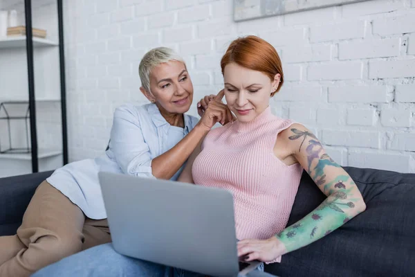 Smiling woman sitting near lesbian girlfriend working on laptop at home — Stock Photo