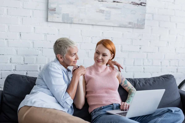 Feliz lésbica mulher abraçando tatuado namorada sentado no sofá com laptop — Fotografia de Stock