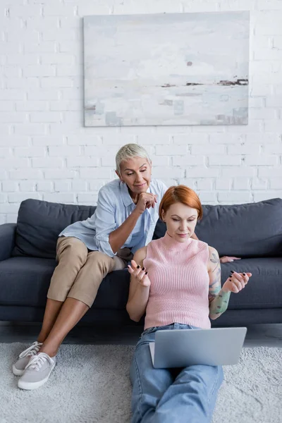 Lesbian woman smiling on couch while discouraged lesbian girlfriend gesturing on floor near laptop — Stock Photo