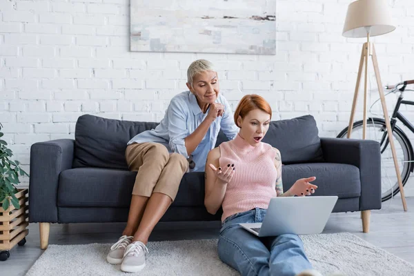 Amazed woman showing wow gesture while sitting on floor with laptop near lesbian girlfriend on couch — Stock Photo