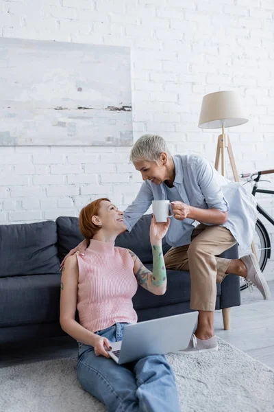 Lesbiana mujer dando taza de té a feliz novia sentado en sofá con portátil - foto de stock