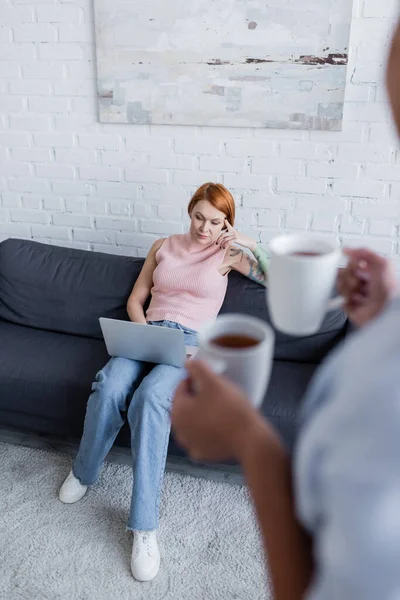 Blurred woman holding cups with tea near thoughtful girlfriend sitting on couch with laptop — Stock Photo