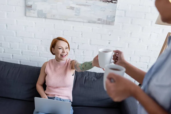 Femme floue tenant des tasses à thé près gai lesbienne copine assis sur canapé avec ordinateur portable — Photo de stock