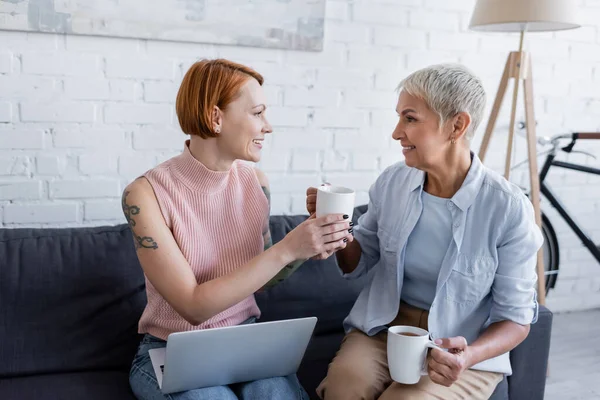 Smiling woman giving cup of tea to tattooed girlfriend sitting on couch with laptop — Stock Photo