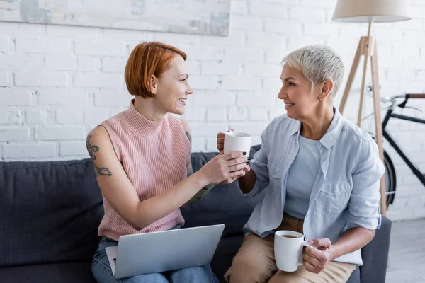 Mujer sonriente dando taza de té a la novia tatuada sentada en el sofá con el ordenador portátil - foto de stock