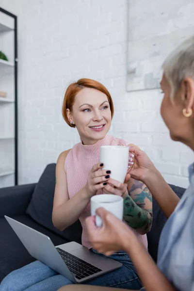 Blurred woman giving tea cup to smiling girlfriend sitting on couch with laptop — Stock Photo