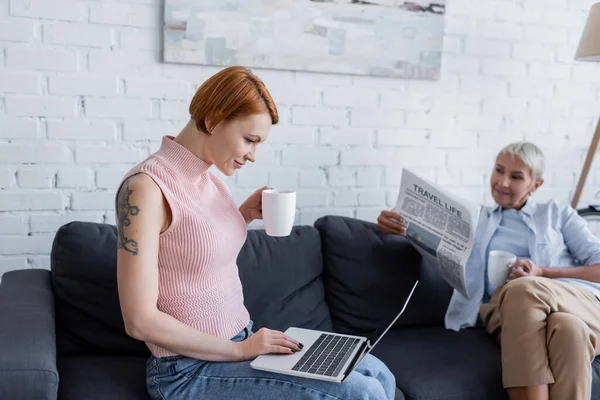 Mujer tatuada con portátil y taza de té cerca de novia borrosa leyendo el periódico de la vida de viaje en la sala de estar - foto de stock