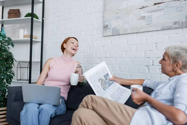 Laughing woman sitting on couch with laptop near lesbian girlfriend with newspaper and cup of tea — Stock Photo