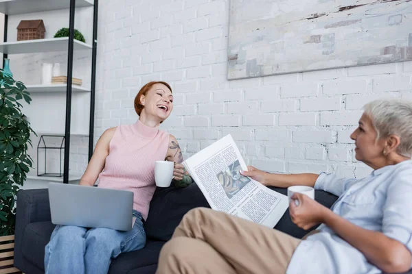 Donna ridente seduta sul divano con computer portatile vicino fidanzata lesbica con giornale e tazza di tè — Foto stock