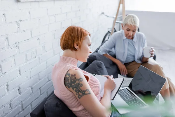 Tattooed woman writing in notebook near laptop with blank screen and blurred lesbian girlfriend with tea cup — Stock Photo