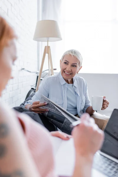 Fröhliche Frau mit Zeitung und Teetasse lächelt auf Couch neben verschwommener Freundin — Stockfoto