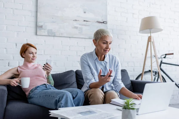 Lesbienne femme travaillant sur ordinateur portable près petite amie assis sur canapé avec smartphone et tasse de thé — Photo de stock