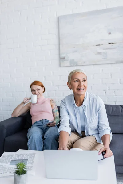 Happy lesbian woman smiling near laptop while blurred girlfriend drinking tea on couch — Stock Photo