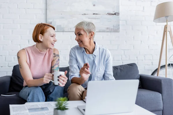 Lesbian woman pointing at laptop near cheerful girlfriend with tea cup — Stock Photo
