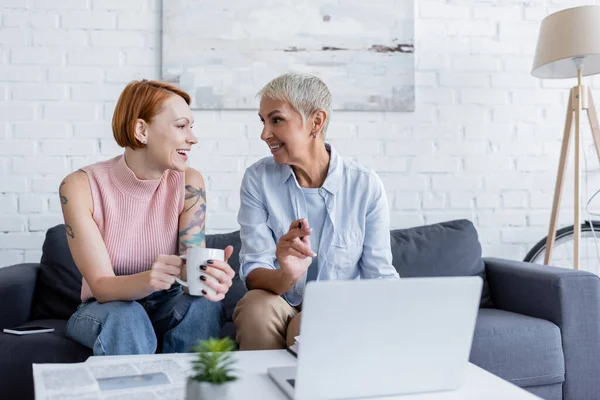 Lesbienne femme pointant vers ordinateur portable près de joyeuse petite amie avec tasse de thé — Photo de stock