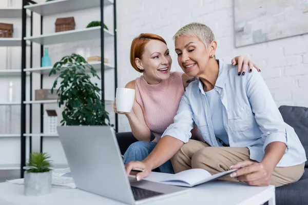 Glückliche Frau mit einer Tasse Tee umarmt lesbische Freundin, die zu Hause am Laptop arbeitet — Stockfoto