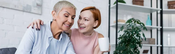 Happy woman with cup of tea talking to lesbian girlfriend at home, banner — Stock Photo
