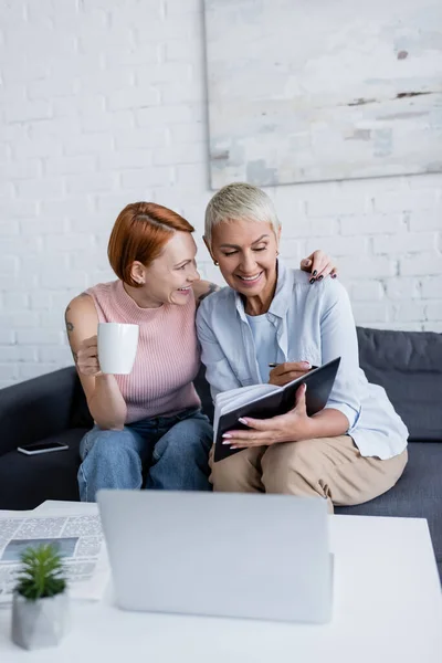 Fröhliche lesbische Frau mit Teetasse umarmt Freundin schreibt in Notizbuch neben verschwommenem Laptop — Stockfoto