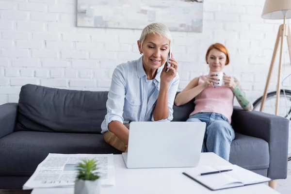 Lächelnde Frau spricht auf Smartphone neben Laptop und lesbische Freundin trinkt Tee auf verschwommenem Hintergrund — Stockfoto