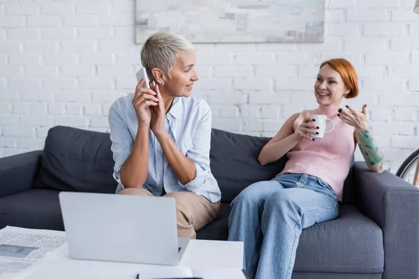 Happy tattooed woman talking to lesbian girlfriend covering smartphone with hand near laptop — Stock Photo