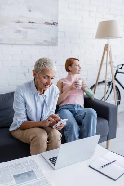 Mujer lesbiana usando teléfono inteligente cerca de la computadora portátil y novia borrosa con taza de té - foto de stock